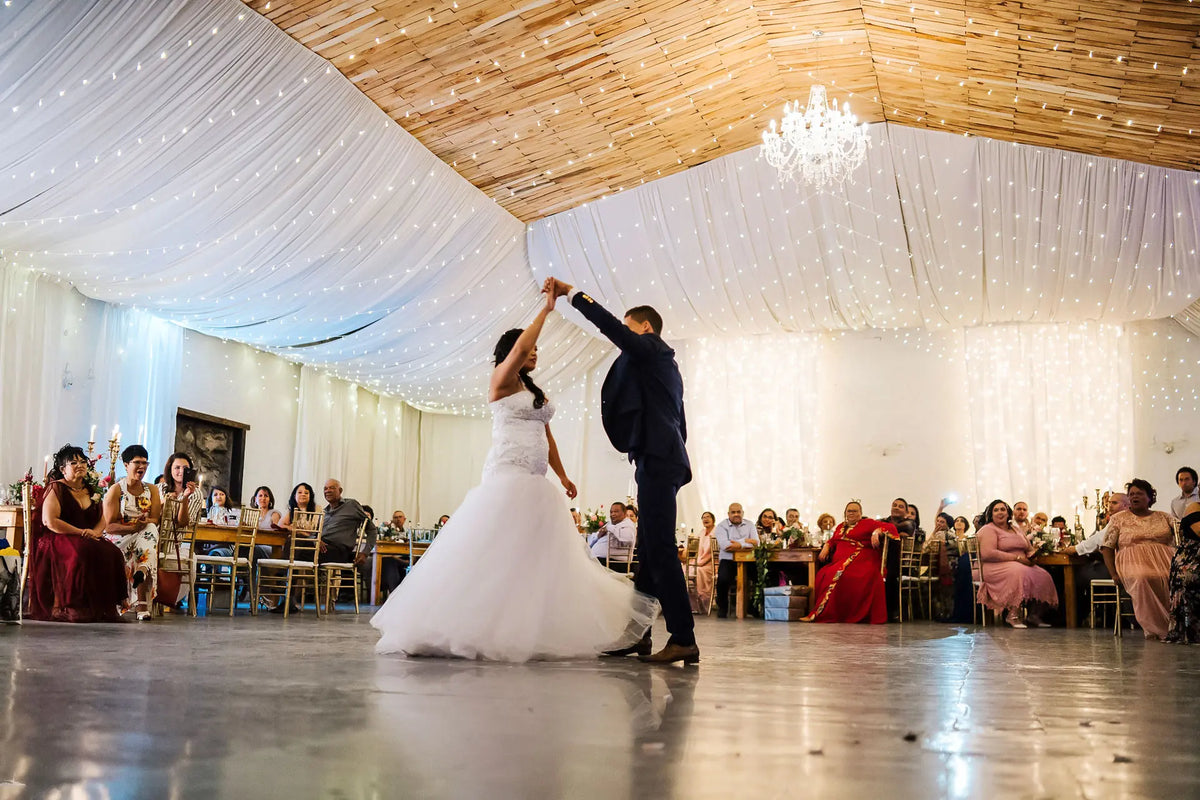 The first dance in the Bezalel Cellar Hall at Bezalel Estate, the hall draped in white beneath a timber ceiling strung with lights and a chandelier, guests at long tables on either side.