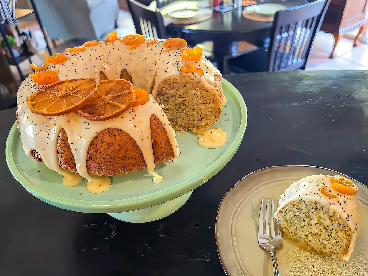 An orange bundt cake with white icing and dried orange slices on a light green cake stand, with a slice served on a plate in the foreground on a dark table.