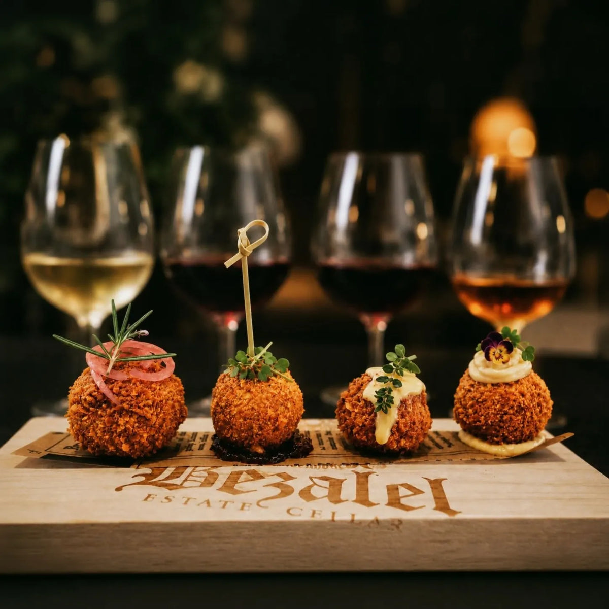 Four appetizer balls served on a branded wooden platter in front of four wine glasses on a dark table. The platter is engraved with 'The Bezalel Estate Cellars'.