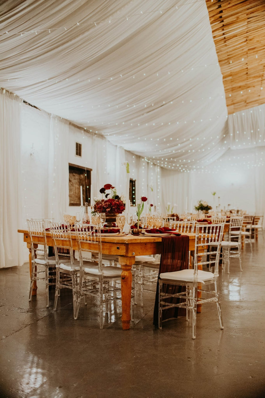 A reception table set with white linen and timber chairs indoors, strung with lights