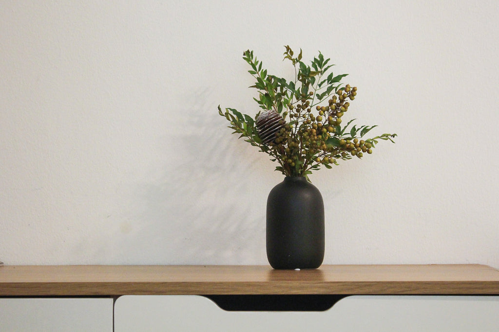 Black vase with greenery on a wooden table against a white wall