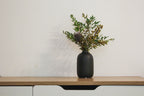 Black vase with greenery on a wooden table against a white wall