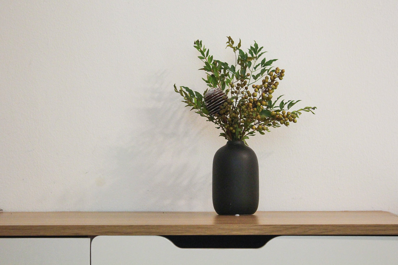 Black vase with greenery on a wooden table against a white wall