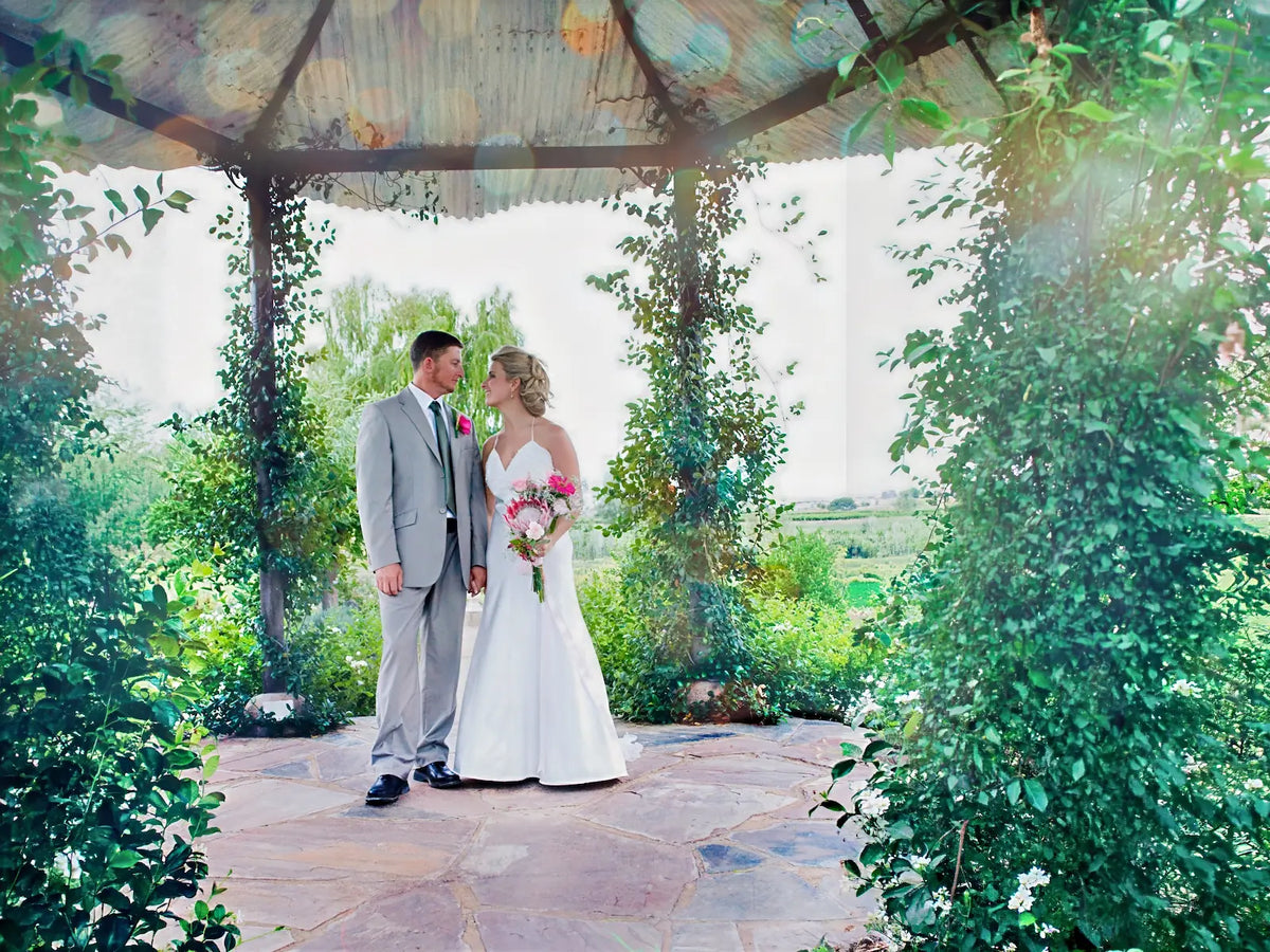 A couple beneath the Pagoda at Bezalel Estate, its timber posts wound with climbing greenery and flowers, the stone floor underfoot and the vineyards showing through the openings.