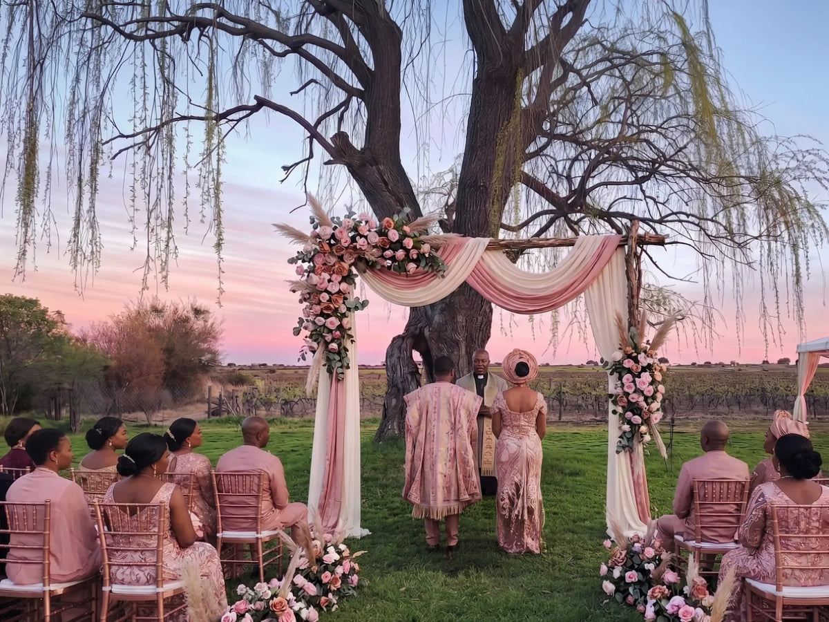 A ceremony beneath the willow at Bezalel Estate: the couple under a floral arch of pale roses and draped fabric, guests seated on the grass, the vineyard rows and a pink dusk sky beyond.